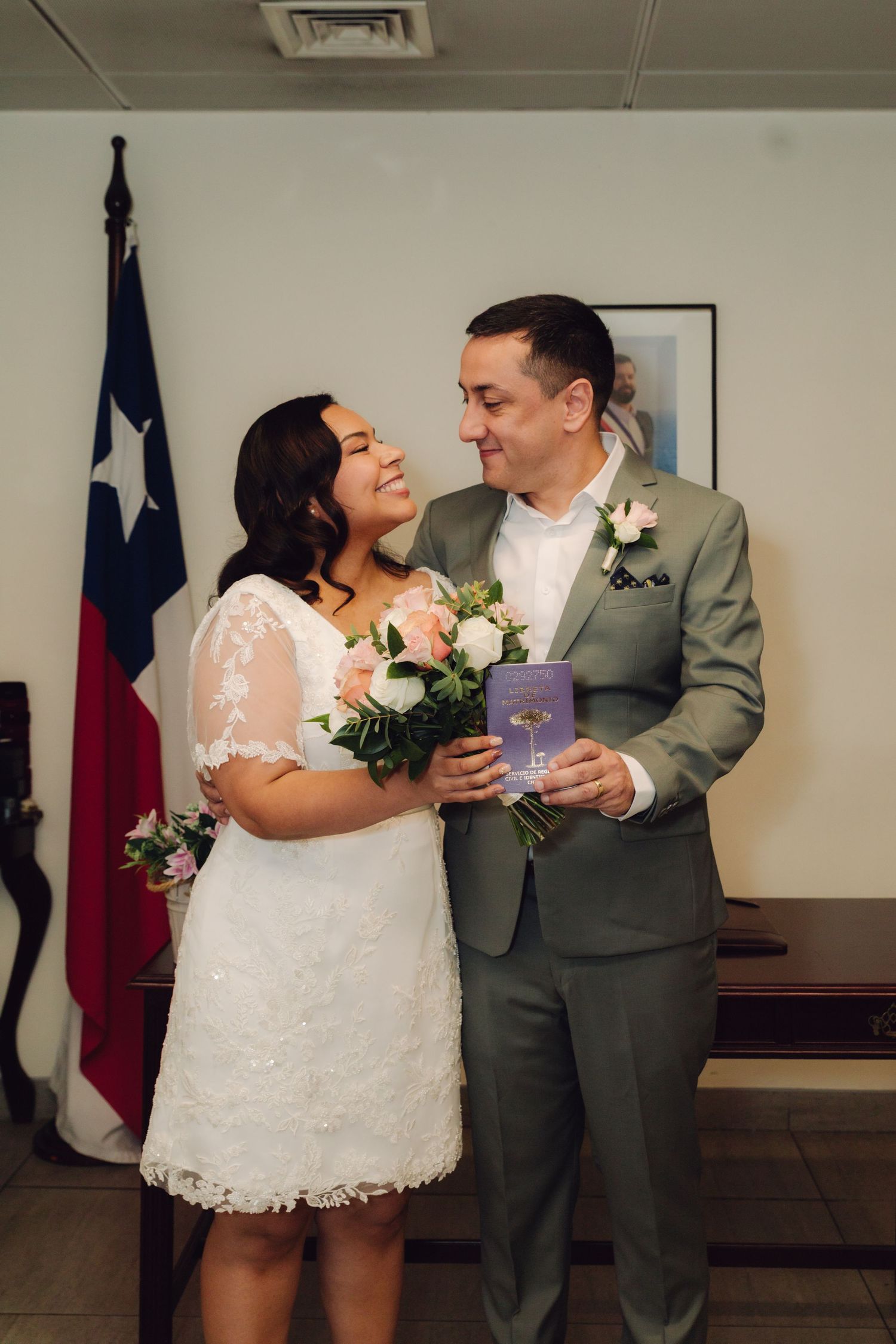Bride and groom share a tender moment holding bouquet and marriage book before Texas flag at courthouse wedding.