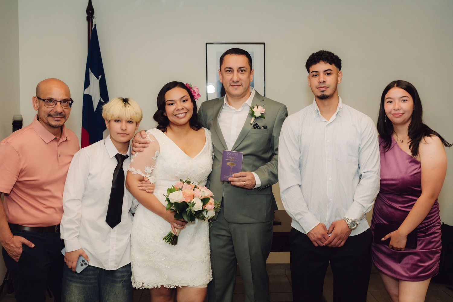 Wedding party group photo with bride in lace dress and groom in gray suit before Texas state flag.
