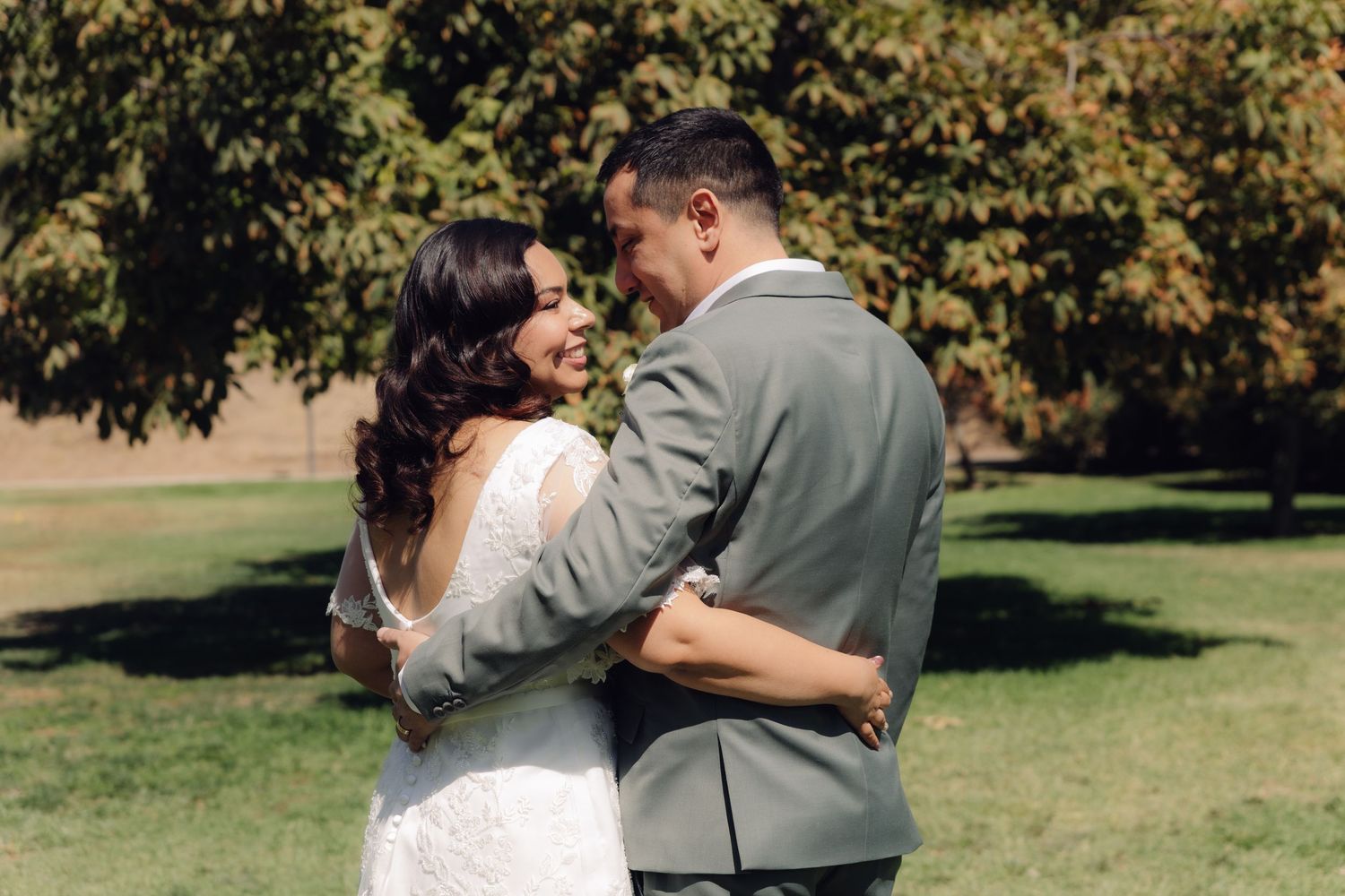 Bride and groom embrace romantically in lush green park setting after outdoor wedding ceremony.