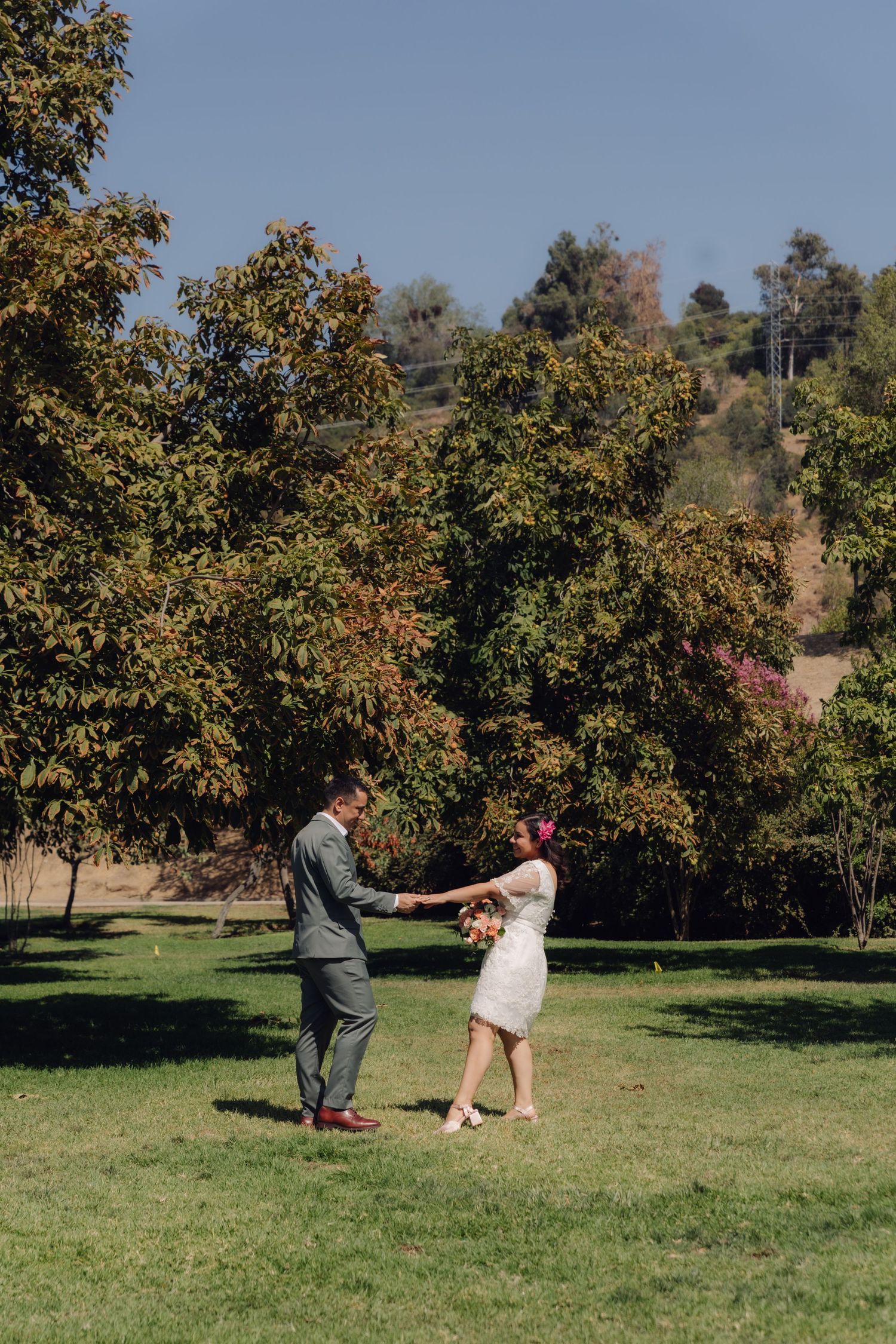 Bride and groom holding hands in a lush green park surrounded by autumn trees on their wedding day.