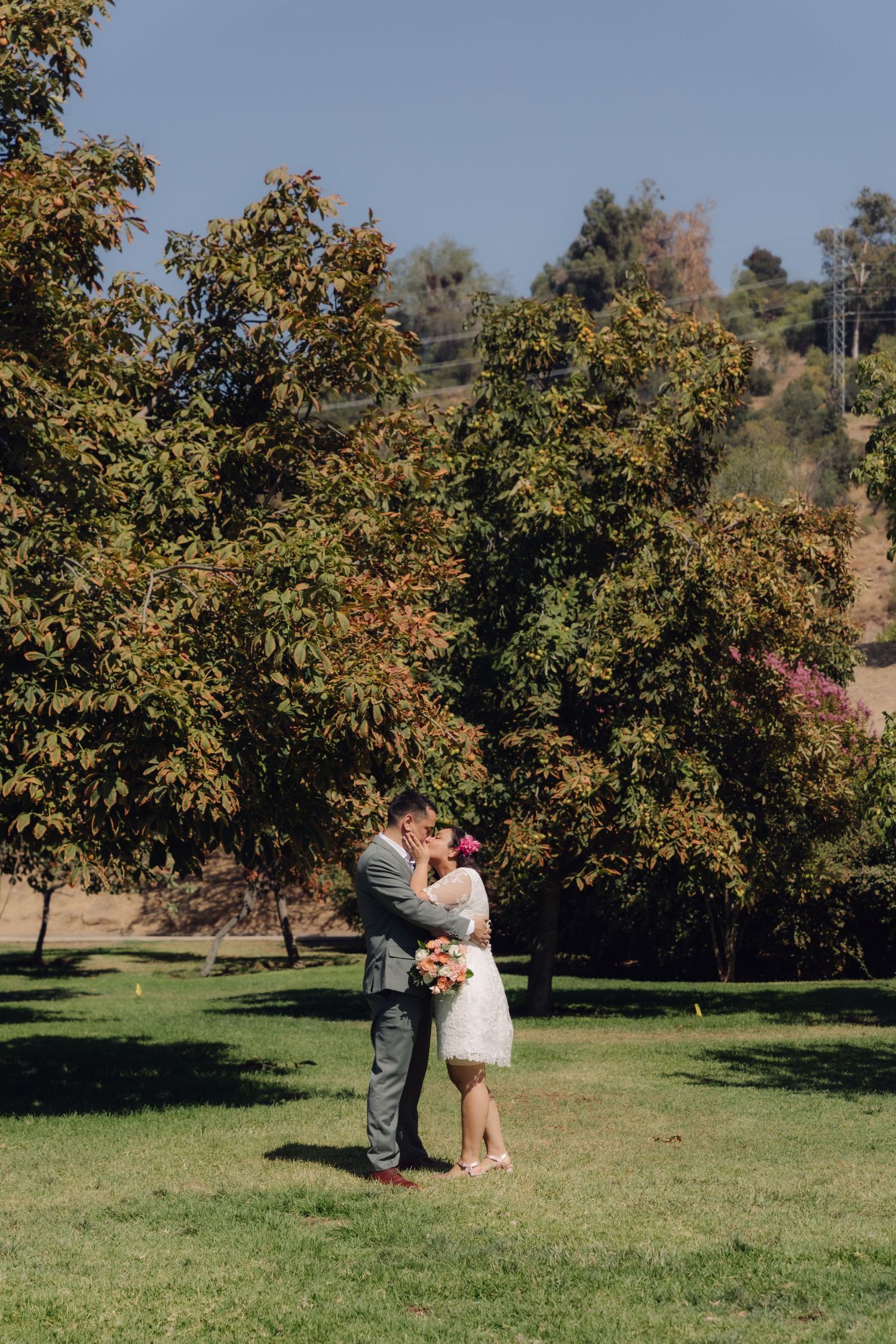Newlyweds share a kiss in scenic park with colorful autumn trees and blue sky backdrop.