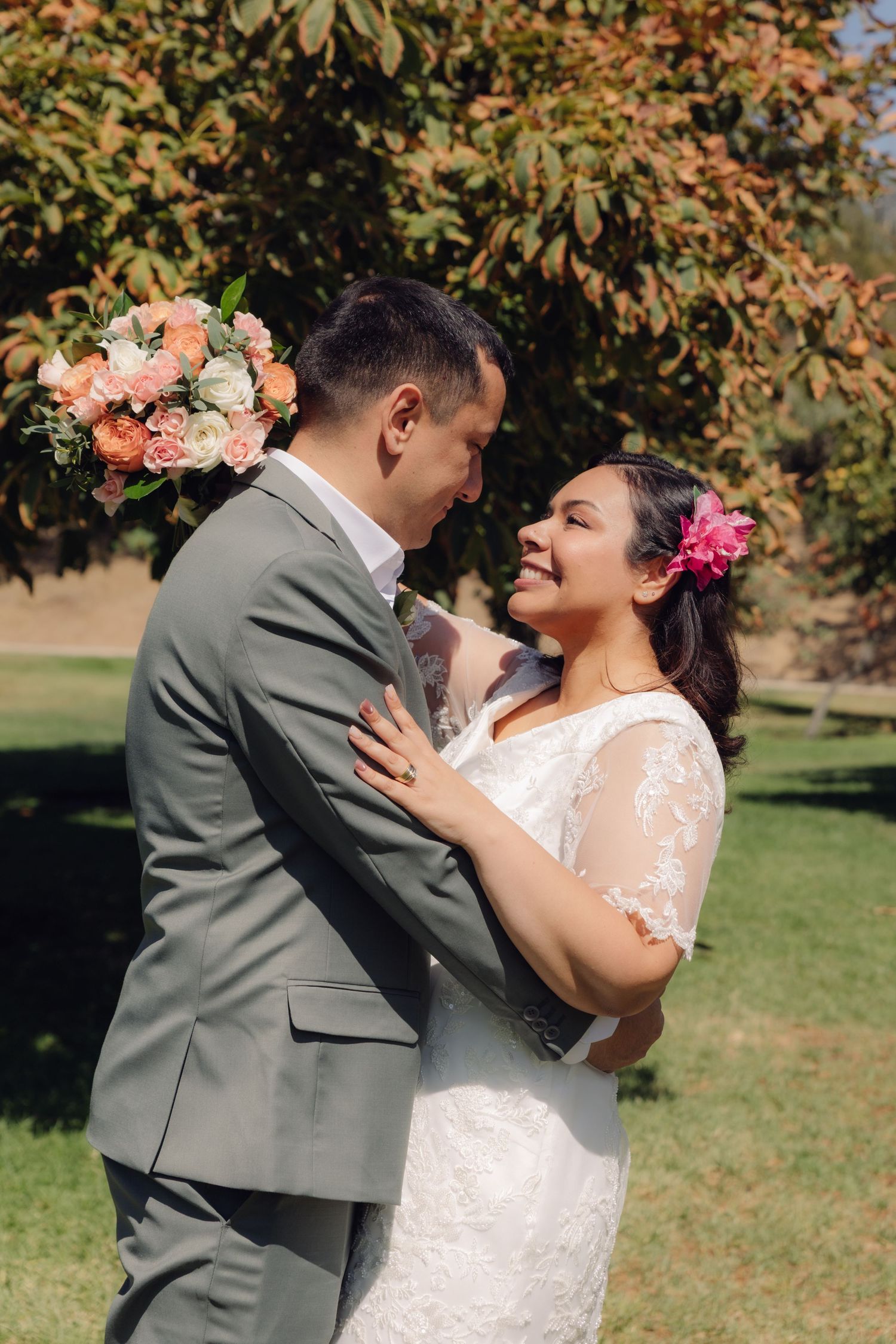 Smiling bride with pink flower in hair embraces groom in gray suit outdoors on wedding day.