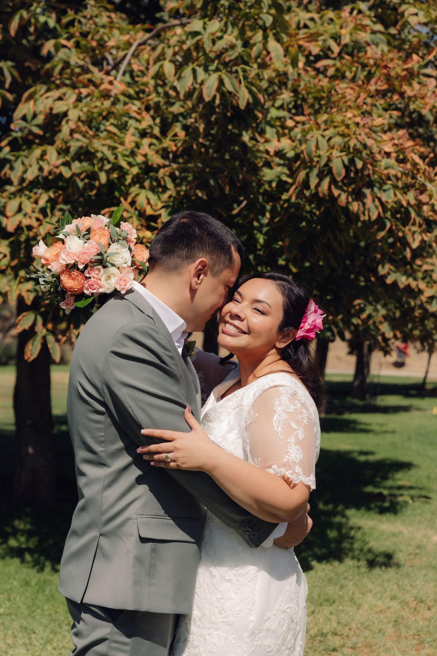 Groom in gray suit kisses bride on cheek as she smiles joyfully holding a peach floral bouquet.
