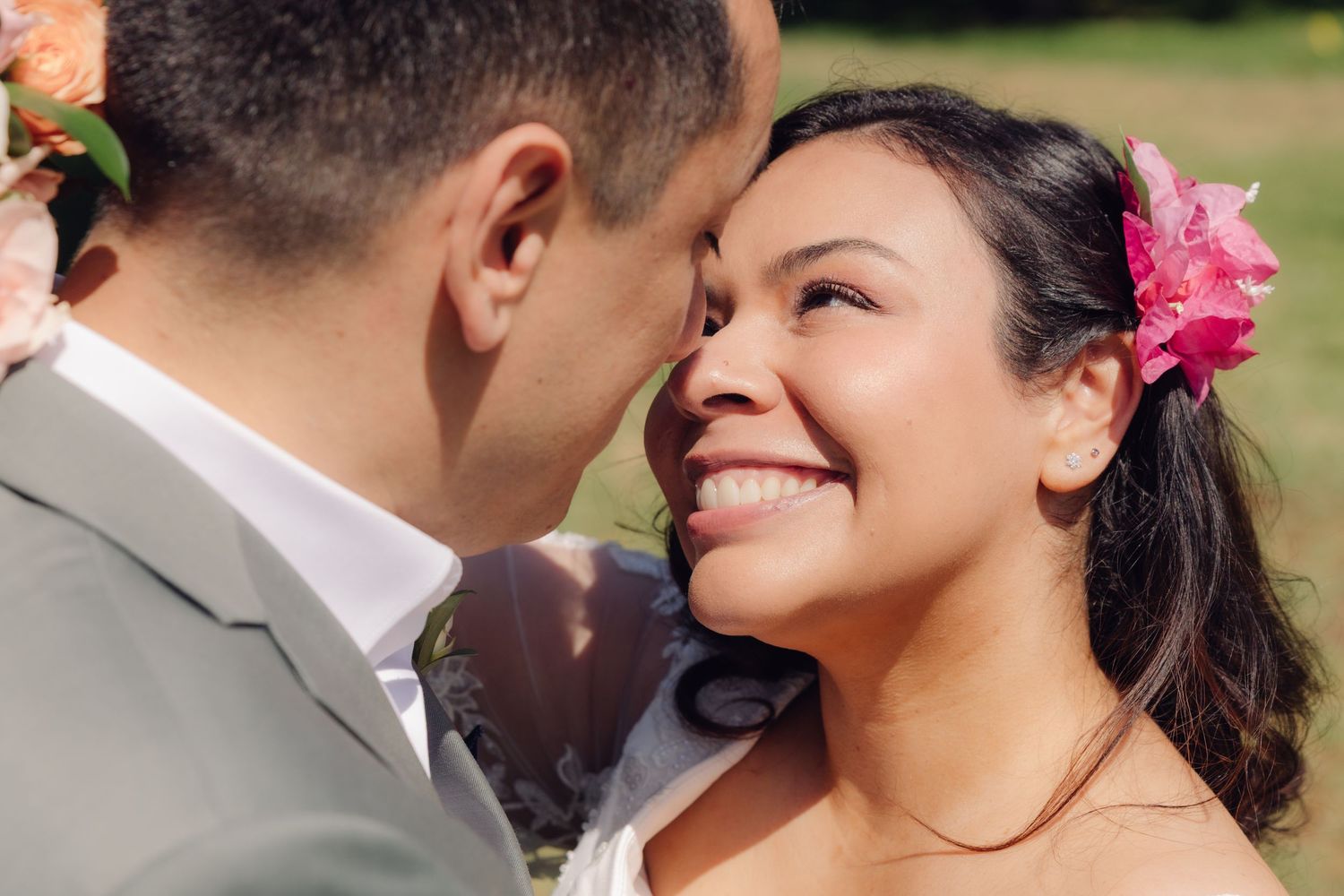 Close-up of radiant bride with pink flower in hair smiling at groom during outdoor wedding shoot.
