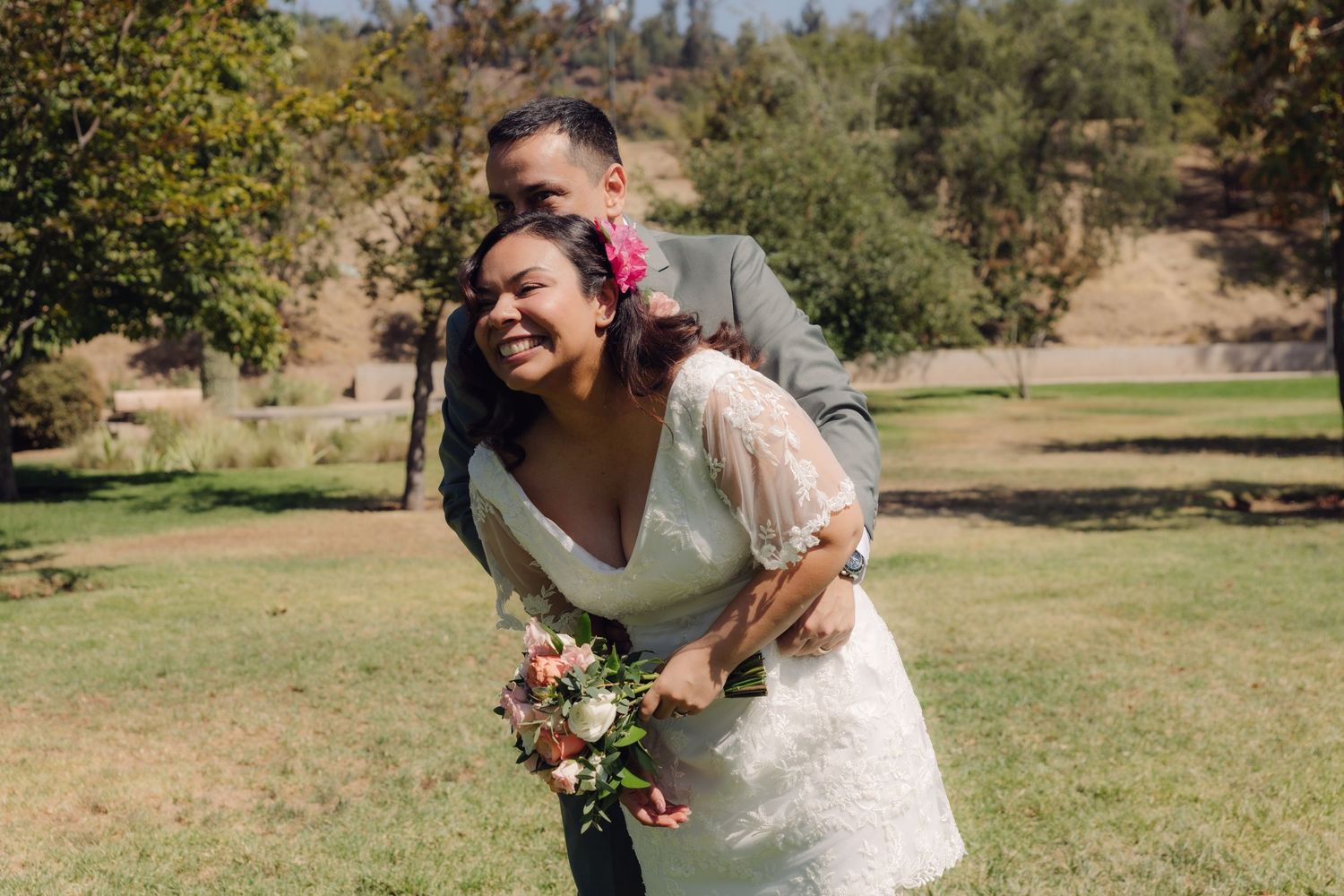 Laughing bride in lace dress leans forward as groom playfully hugs her from behind in a park.