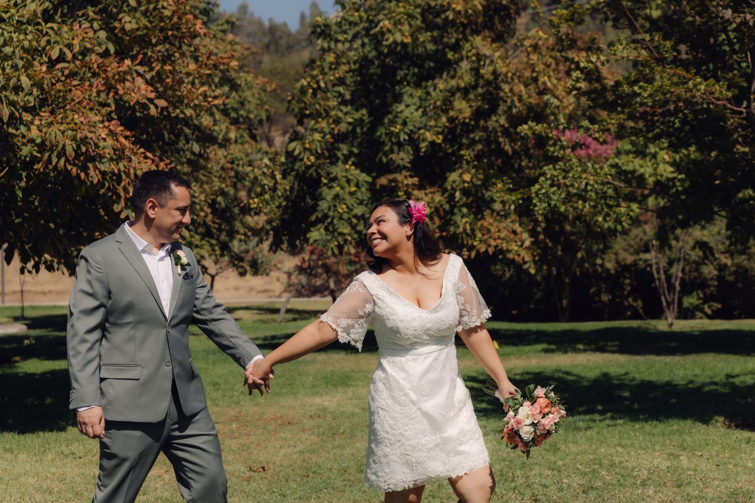 Happy bride and groom walk hand in hand through a sunny park with autumn foliage in background.