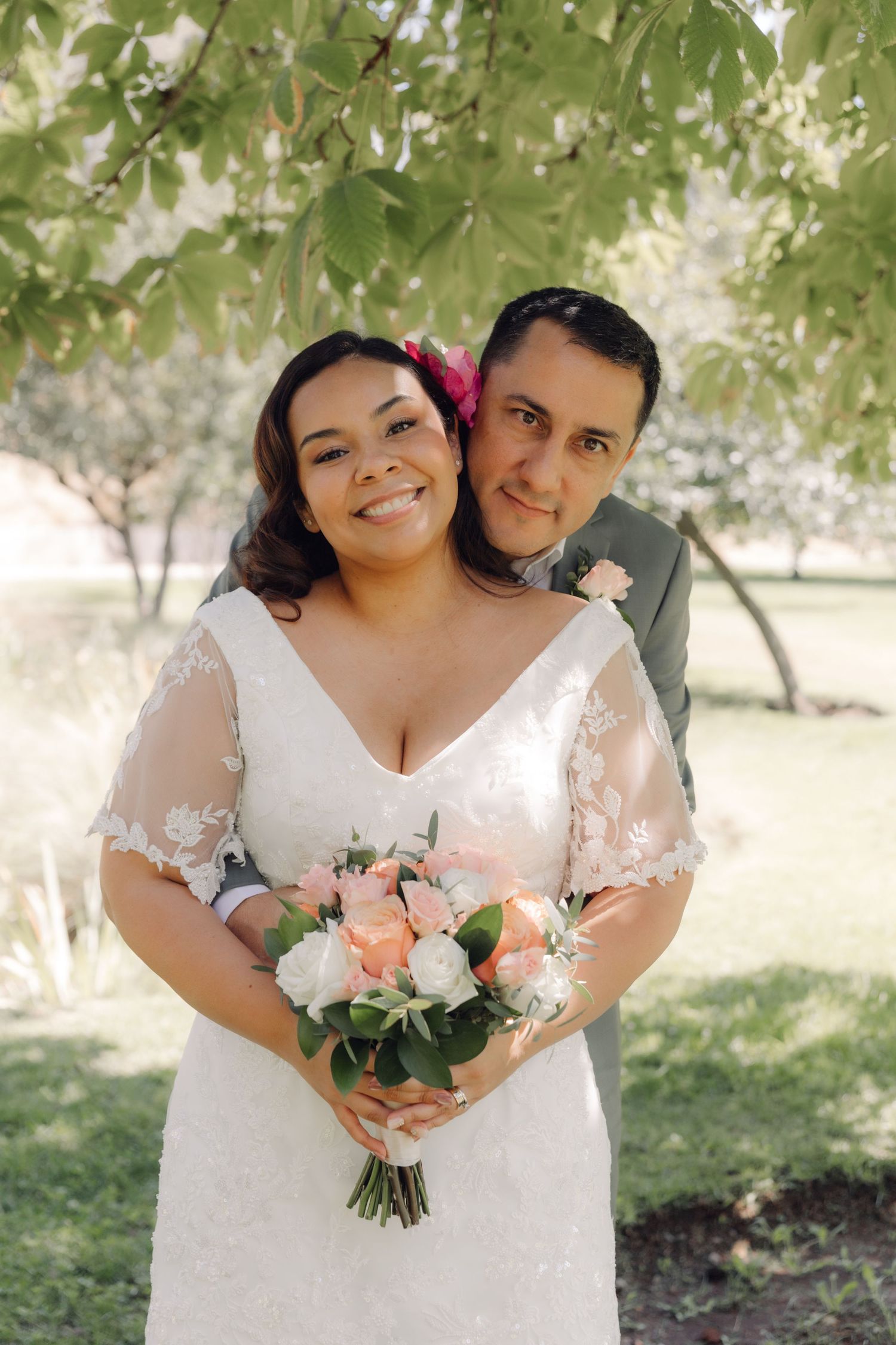 Groom embraces bride from behind as she smiles holding a pink and white rose bouquet under a tree.