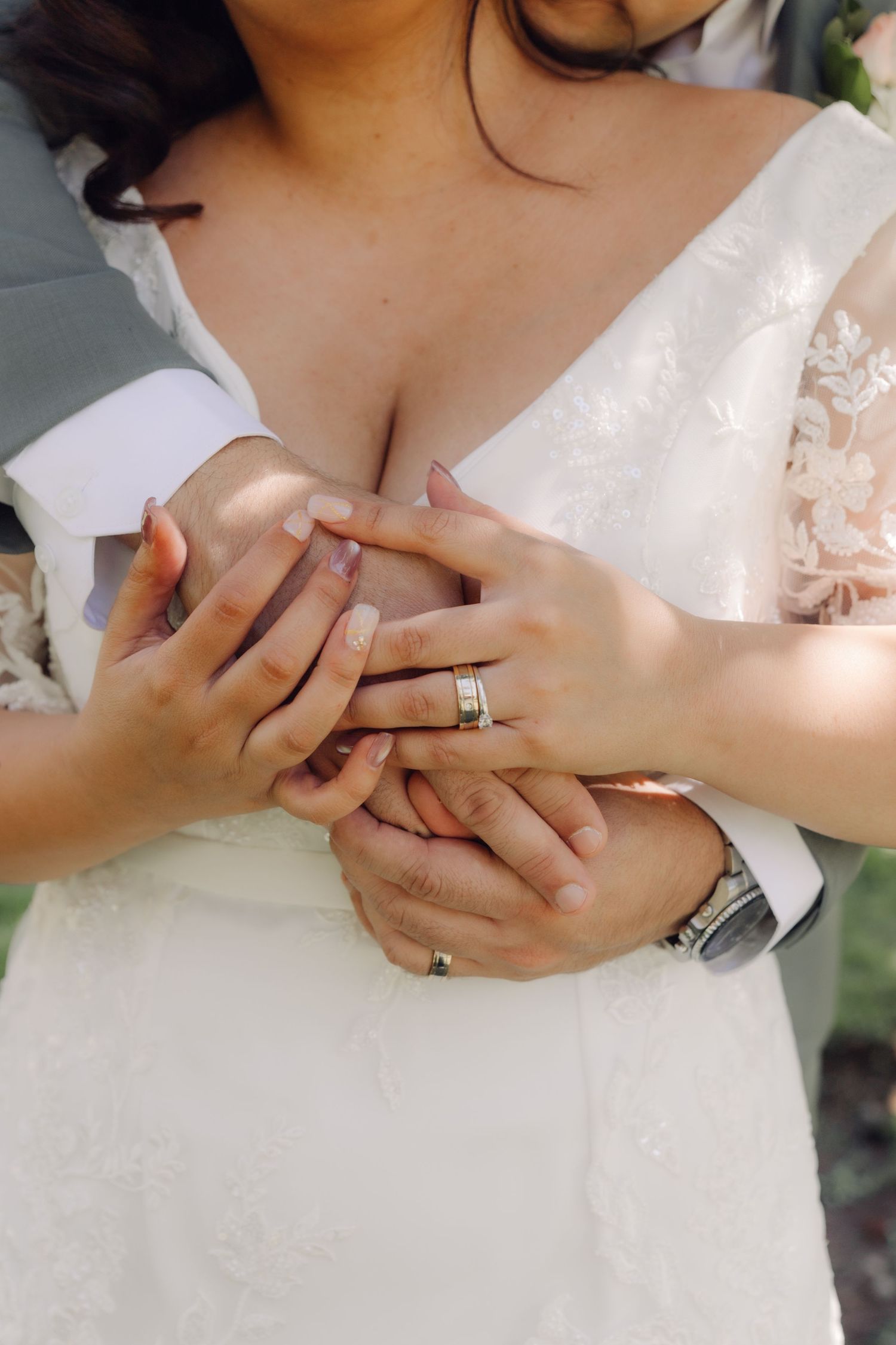 Close-up of bride and groom clasping hands together showing wedding rings against lace bridal gown.
