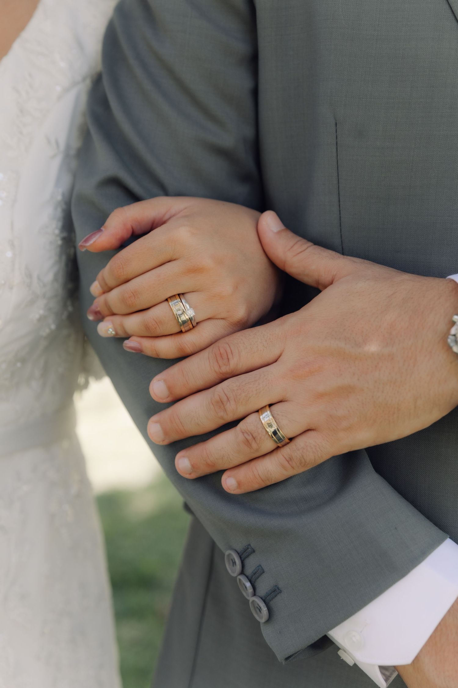 Close-up of bride and groom displaying matching gold wedding bands on their hands against gray suit.