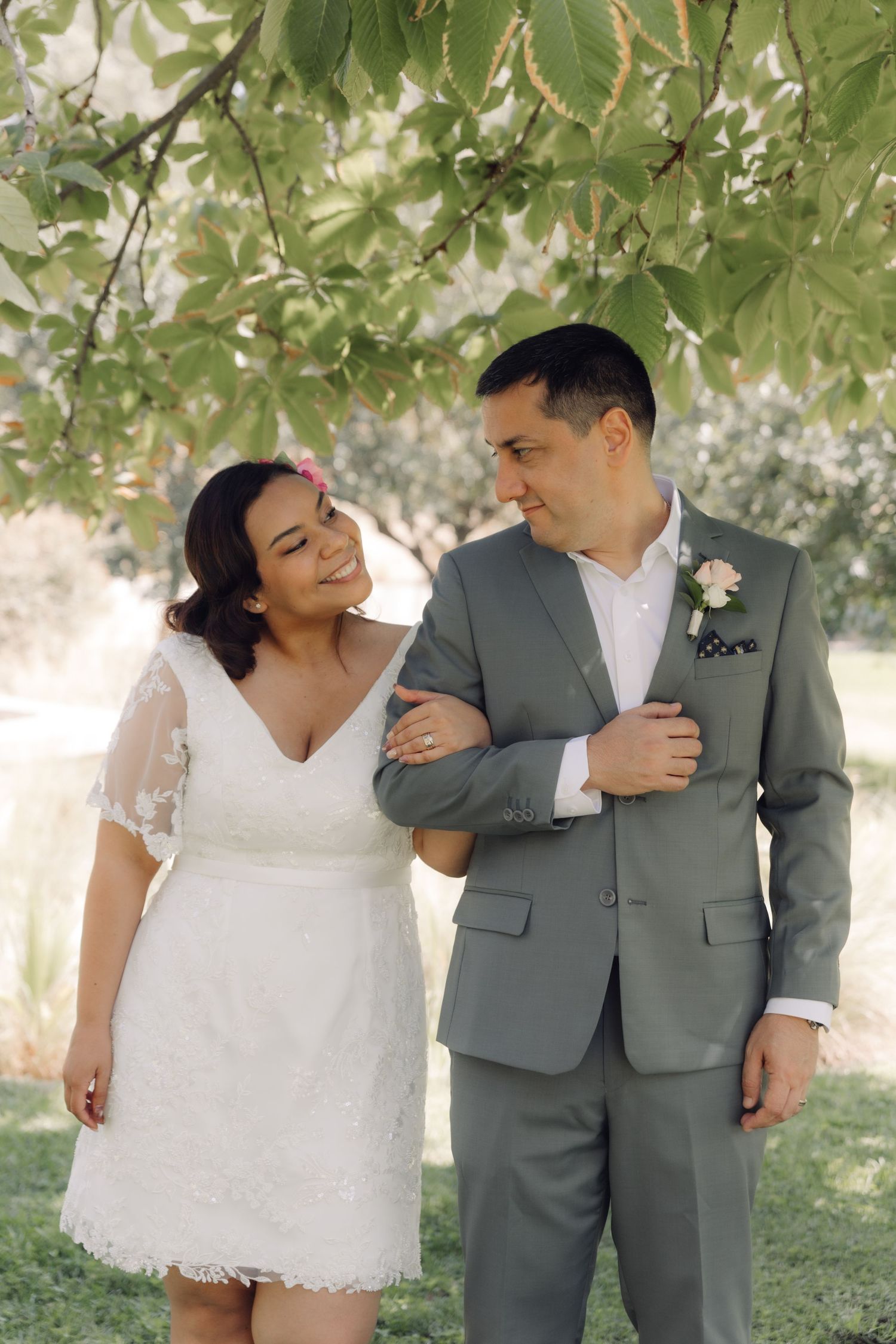 Bride in white lace dress and groom in gray suit share a loving glance under green tree branches outdoors.