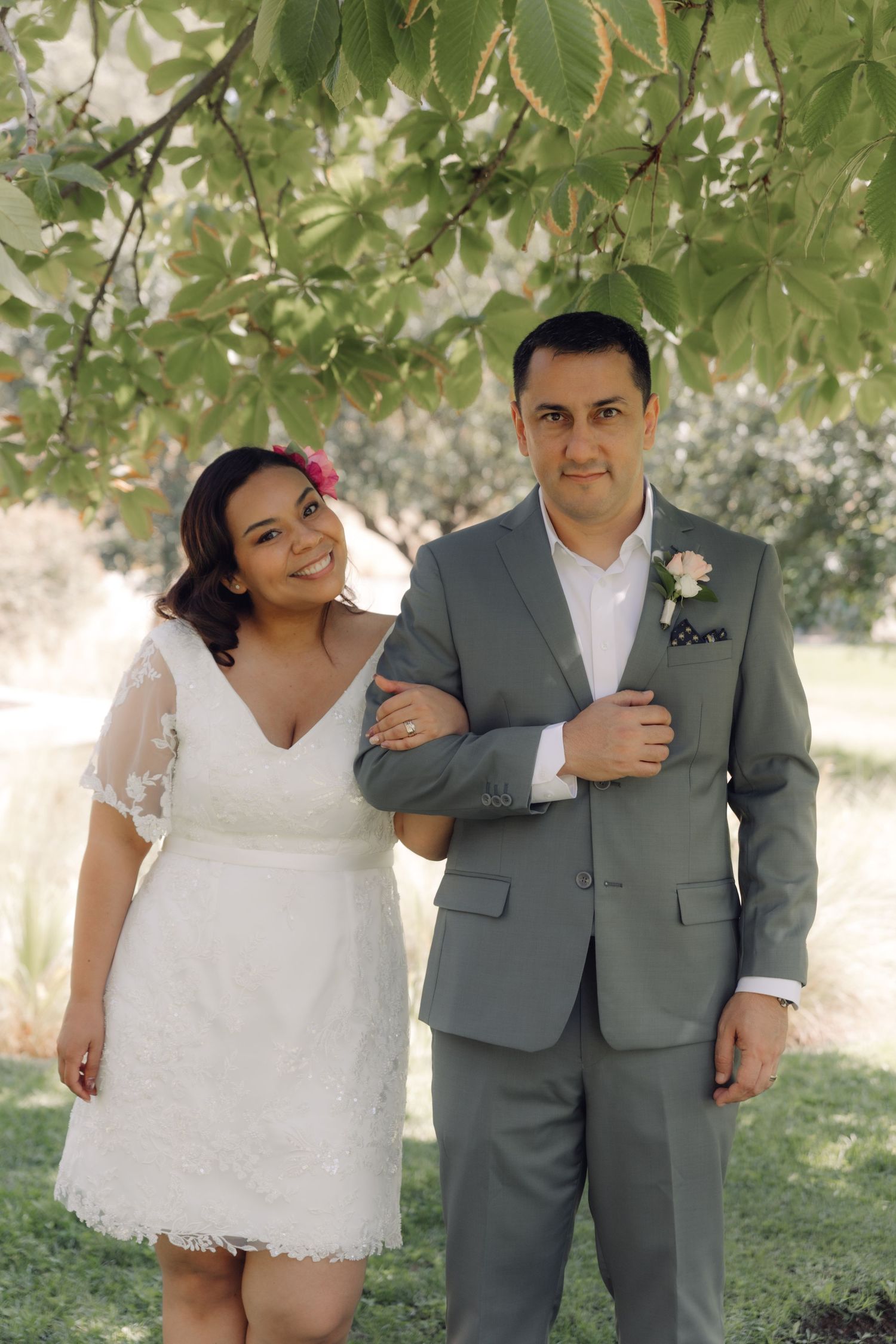 Smiling bride in white lace dress holds grooms arm as they pose together under leafy tree branches.