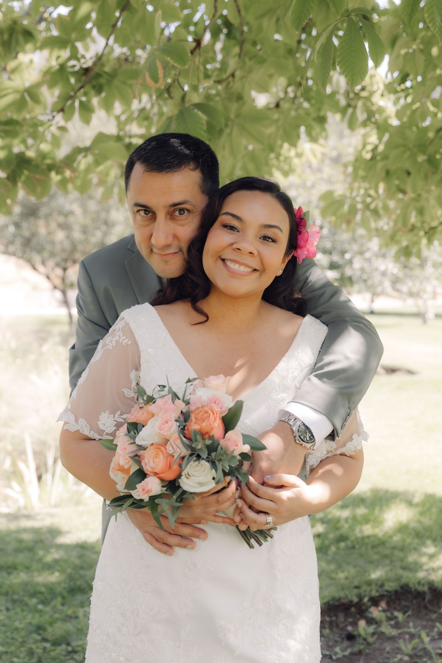 Groom in gray suit embraces bride holding pink and white bouquet under lush green tree branches.