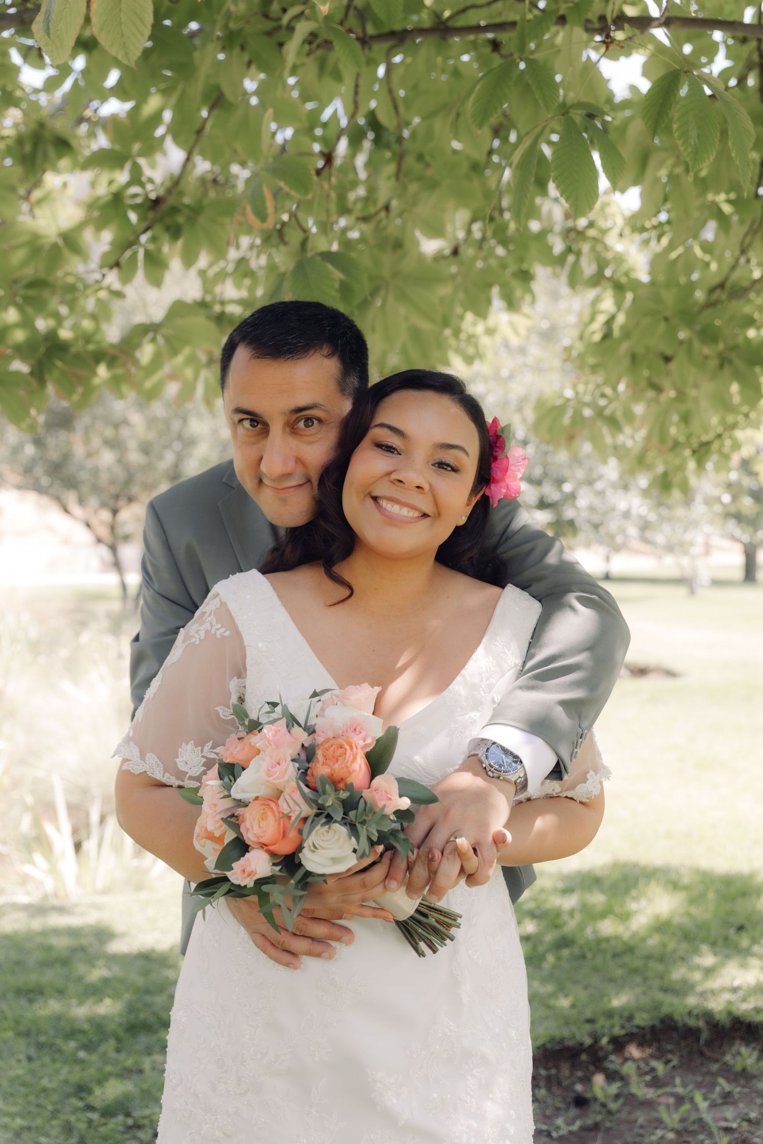 Happy couple poses under green tree, bride holding peach rose bouquet as groom wraps arms around her.