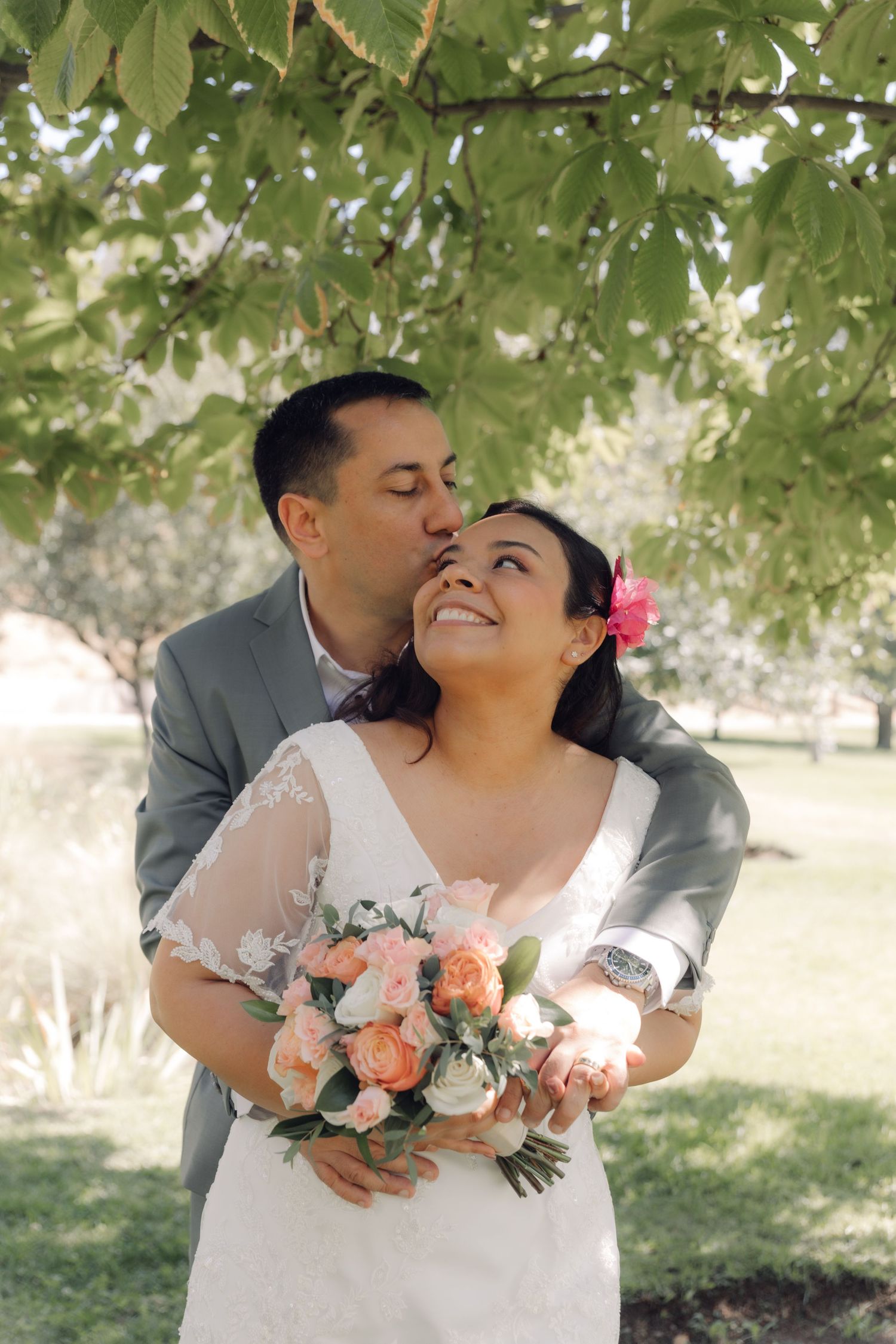 Groom in gray suit kisses brides cheek as she smiles holding a peach and white floral bouquet.