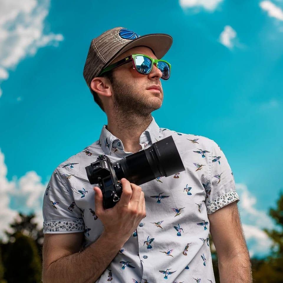 A young man holding a camera looks up wearing sunglasses and a cap against a bright blue sky.