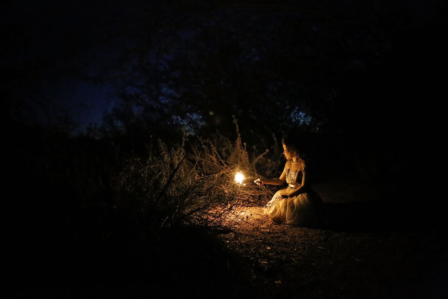 Person in a long dress sits beside a glowing lantern in a dark forest at night, lit by soft warm light.