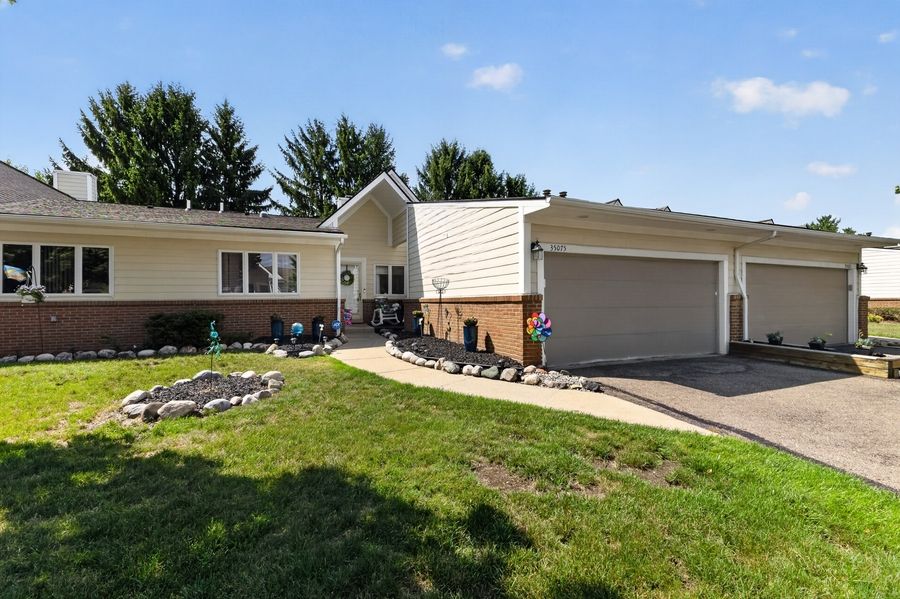 Ranch-style home with attached two-car garage, manicured lawn, decorative rock landscaping, and mature trees on a sunny day.