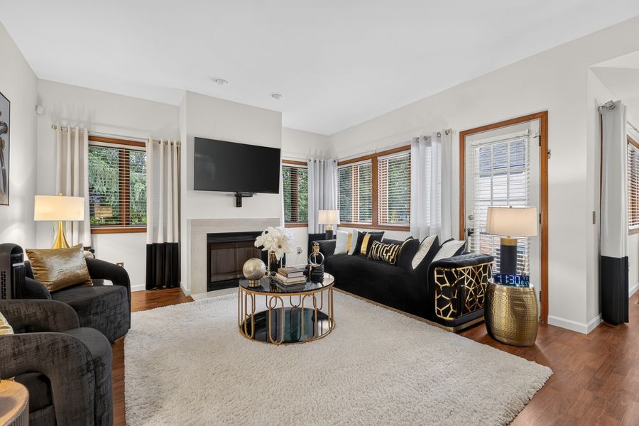 Elegant living room with black sofas, gold accents, fireplace, wall-mounted TV, and large windows with white curtains.