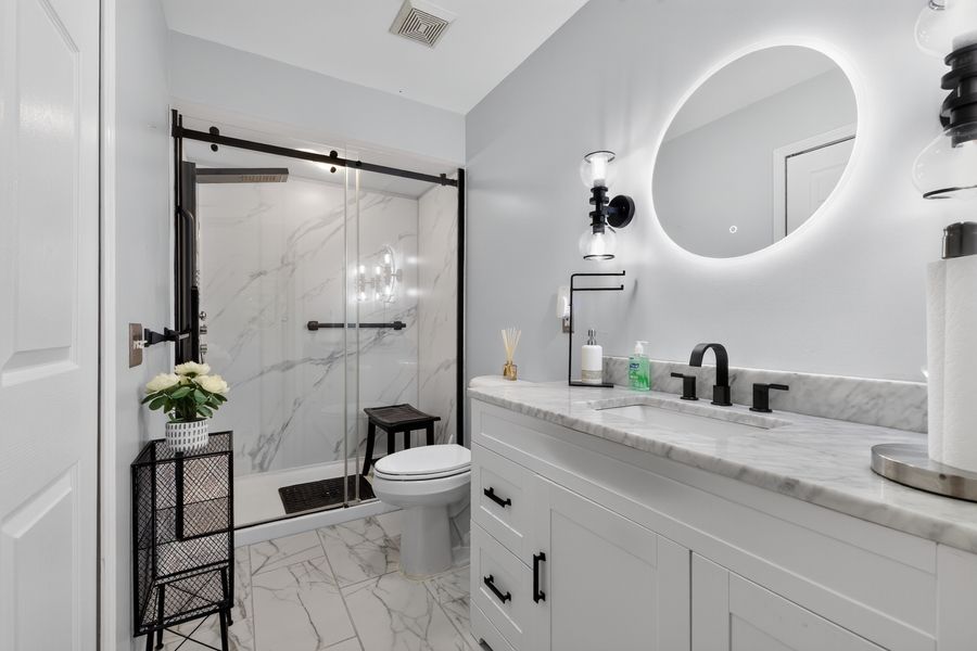 Modern white bathroom with marble shower, round LED mirror, black fixtures, and double vanity.