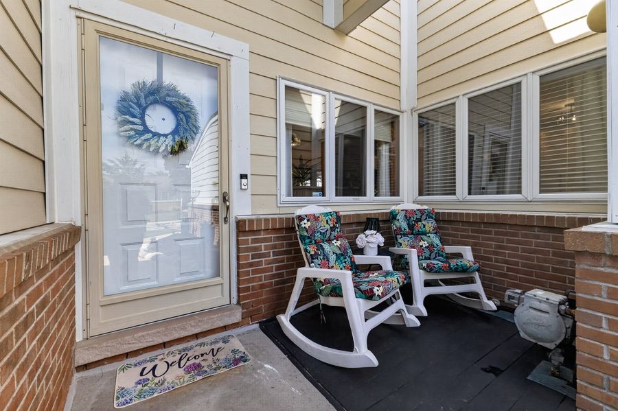 Cozy front porch with two floral rocking chairs, a wreath-adorned white door, and a Welcome mat on brick flooring.