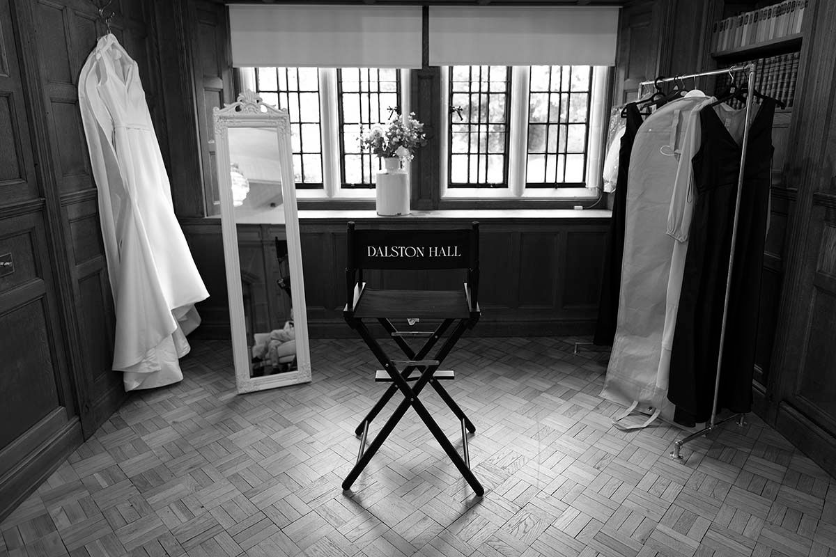 Black and white photo of Dalston Hall director chair between wedding dress and garments on rack.