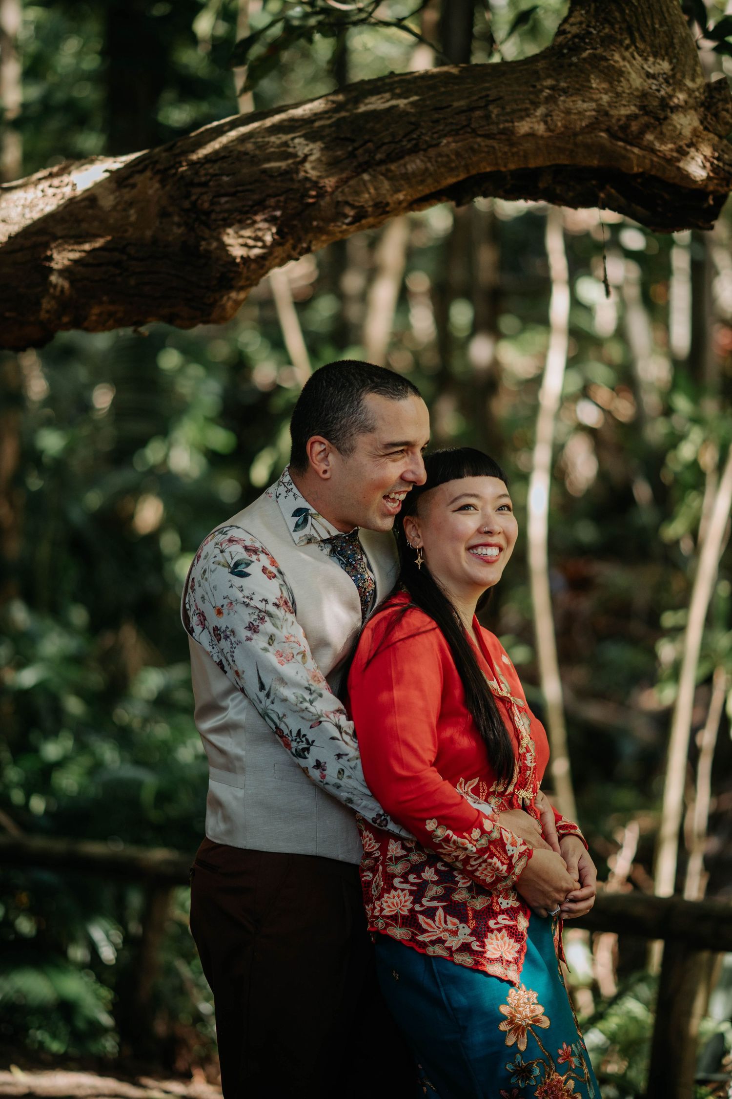 Couple in traditional Asian wedding attire smiling together in a lush forest setting.