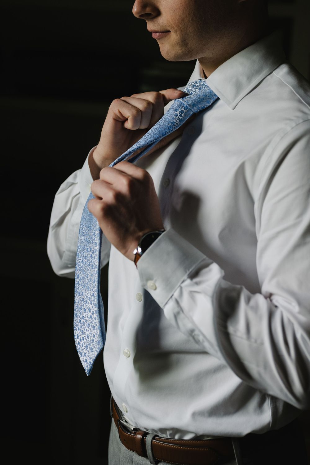 Groom adjusting blue patterned tie over white dress shirt, wearing a brown leather belt and watch.