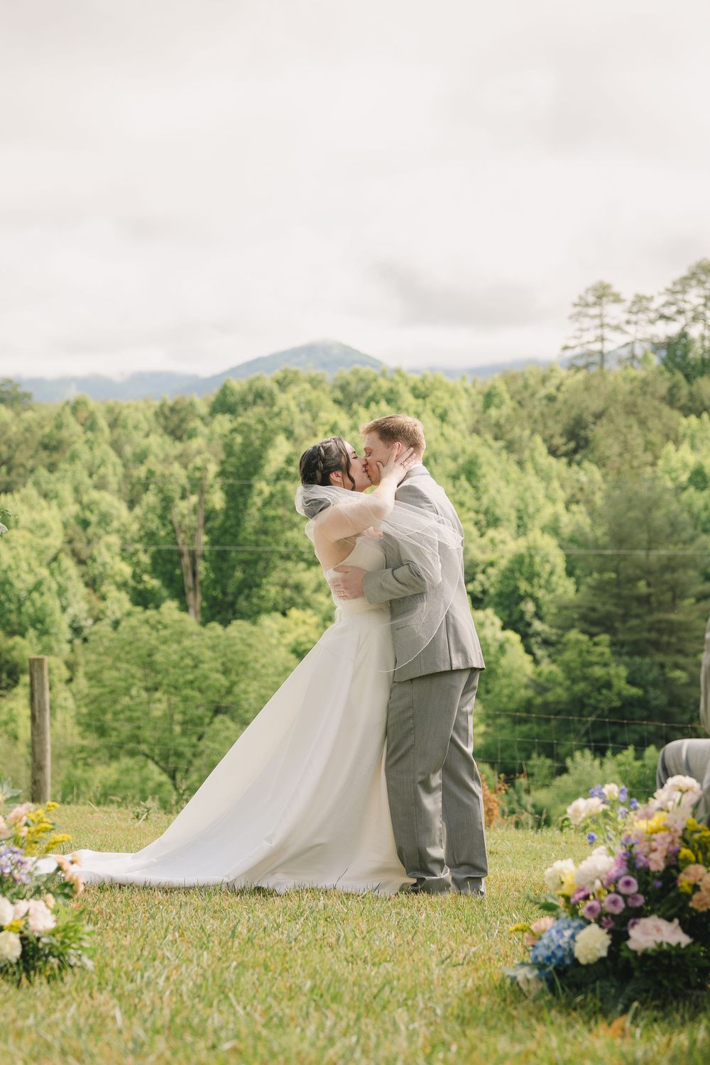 Bride and groom share first kiss at outdoor wedding ceremony with lush green mountain forest backdrop.