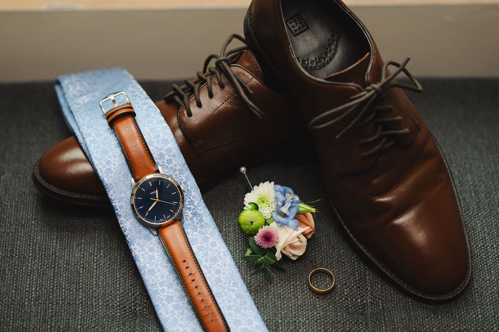 Brown leather dress shoes, a navy watch, blue tie, gold ring, and floral boutonniere arranged for a wedding day.