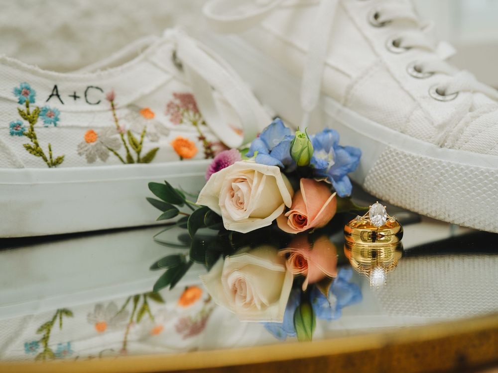 Custom embroidered white wedding sneakers with floral design and gold rings beside colorful boutonniere flowers on reflective surface.