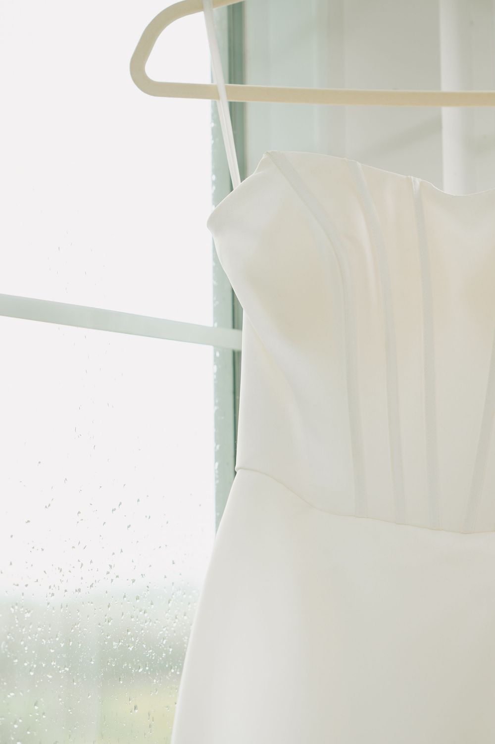 White strapless wedding dress hanging on a wooden hanger by a rain-speckled window with soft natural light.