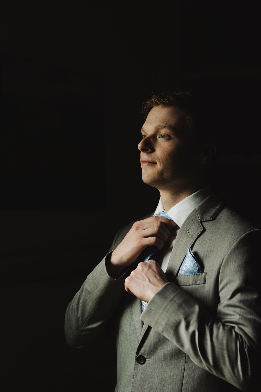 Groom in gray suit adjusting blue tie against dark background, elegant wedding portrait.