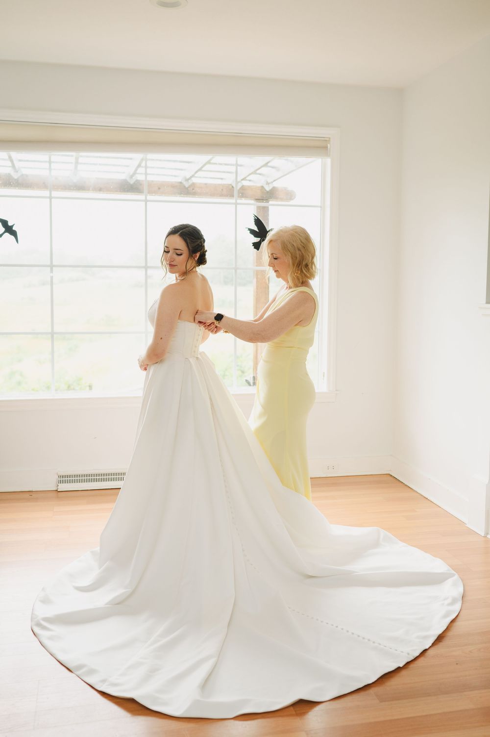 Bride in white gown being helped by bridesmaid in yellow dress near bright window on wedding day.