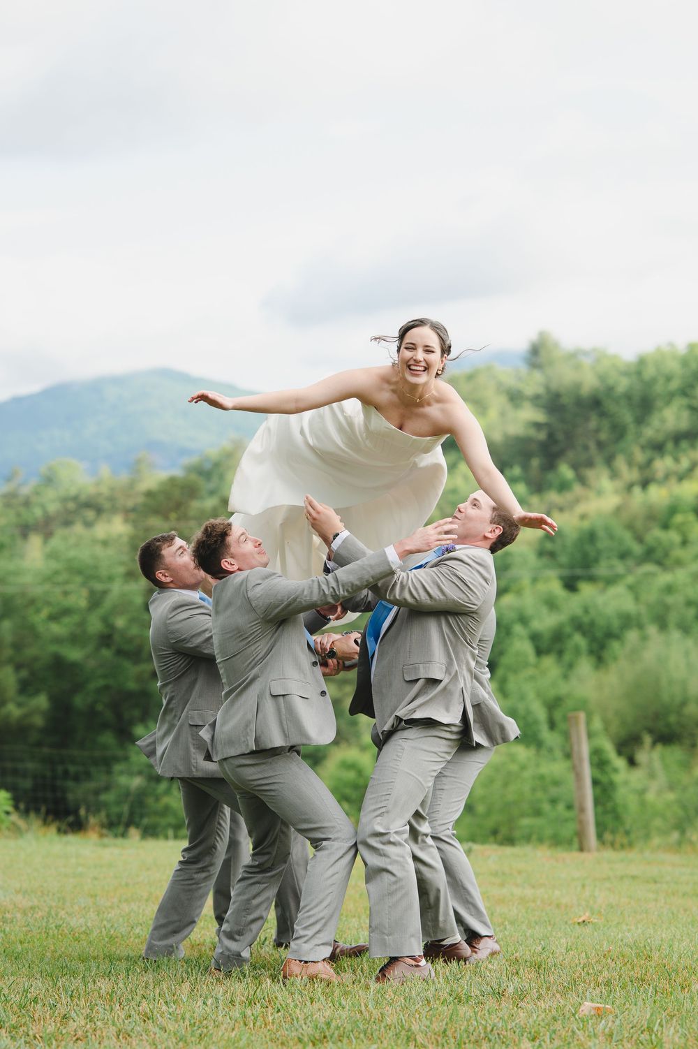 Groomsmen in gray suits tossing a smiling bride in a white dress into the air outdoors with mountains in the background.