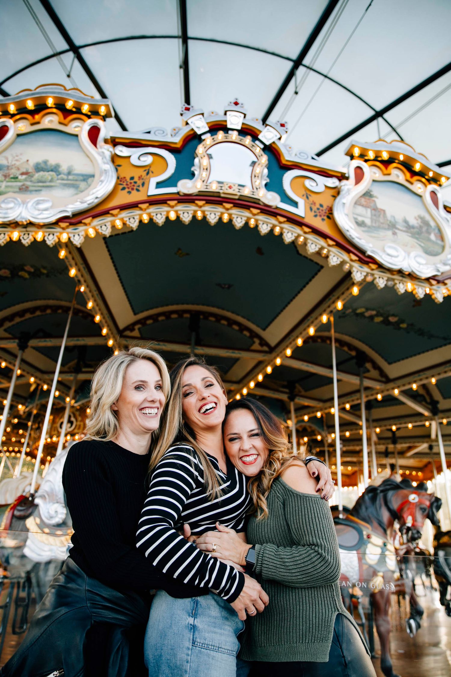 Three friends laugh together while enjoying a vintage carousel ride at an amusement park.
