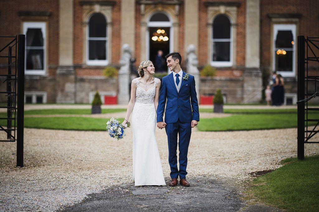 Bride and groom holding hands outside a grand red-brick manor house on their wedding day.