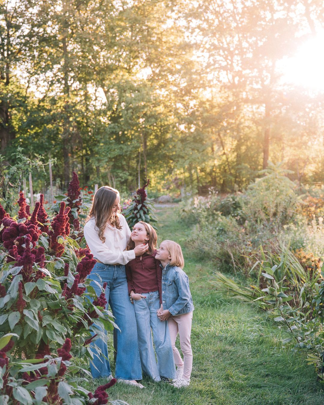 A smiling woman in a denim jacket receives kisses from two young girls outdoors in a lush green setting.
