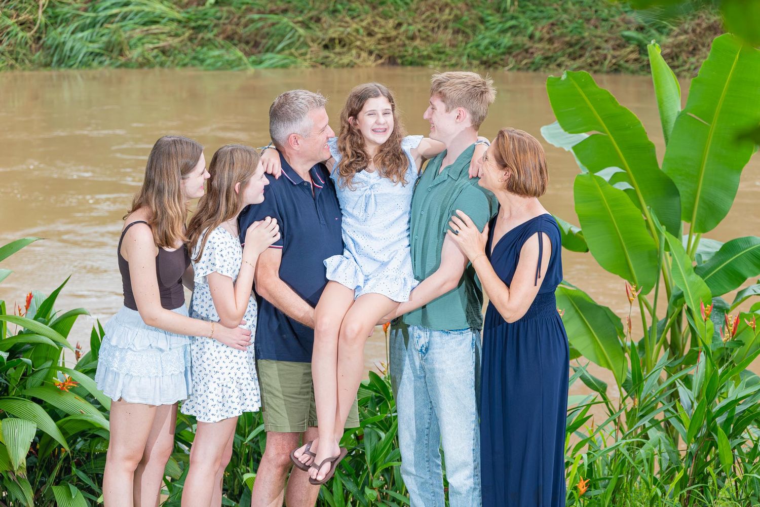 Family of six posing together outdoors near a river surrounded by lush tropical greenery.