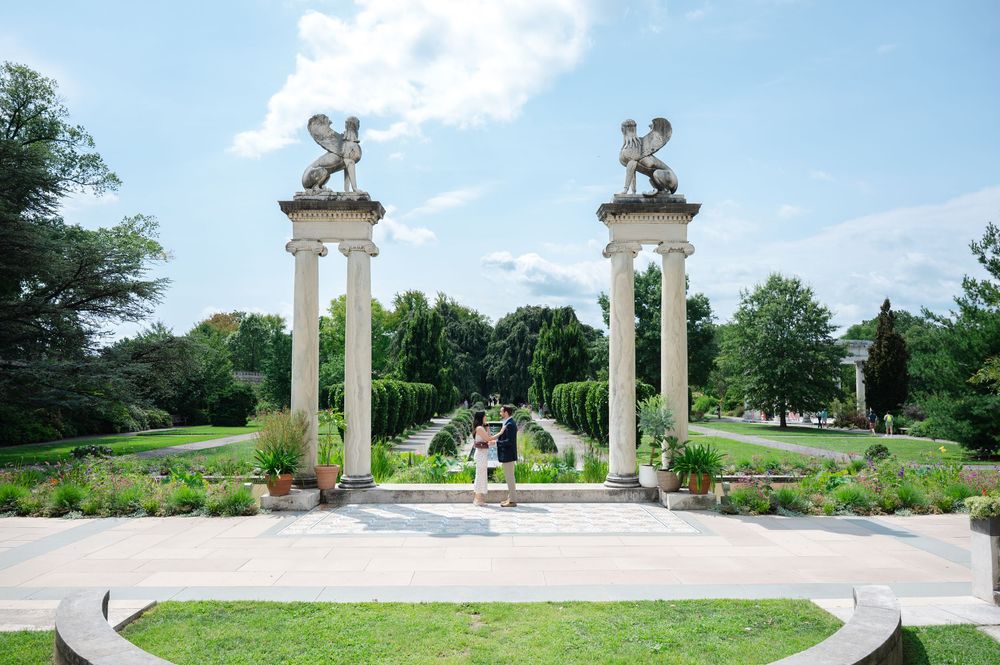 Elegant garden with tall columns topped by griffin statues framing a lush green pathway under a blue sky.