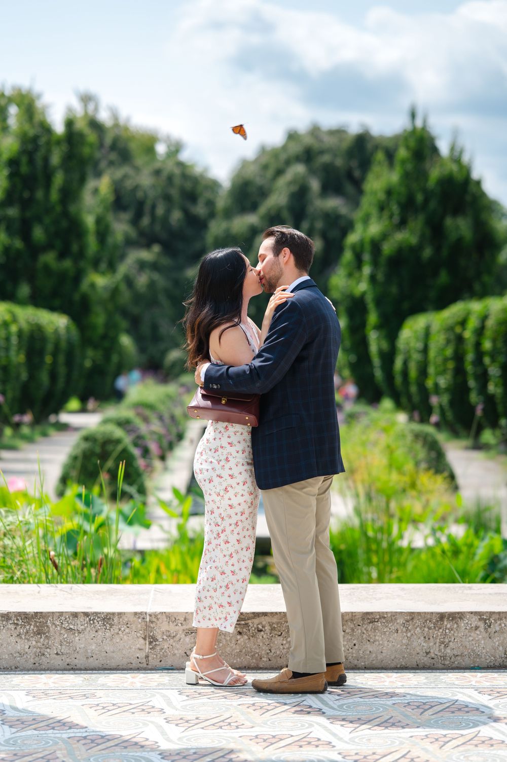 Couple sharing a romantic kiss in a lush garden park, woman in floral dress, man in navy blazer on a sunny day.
