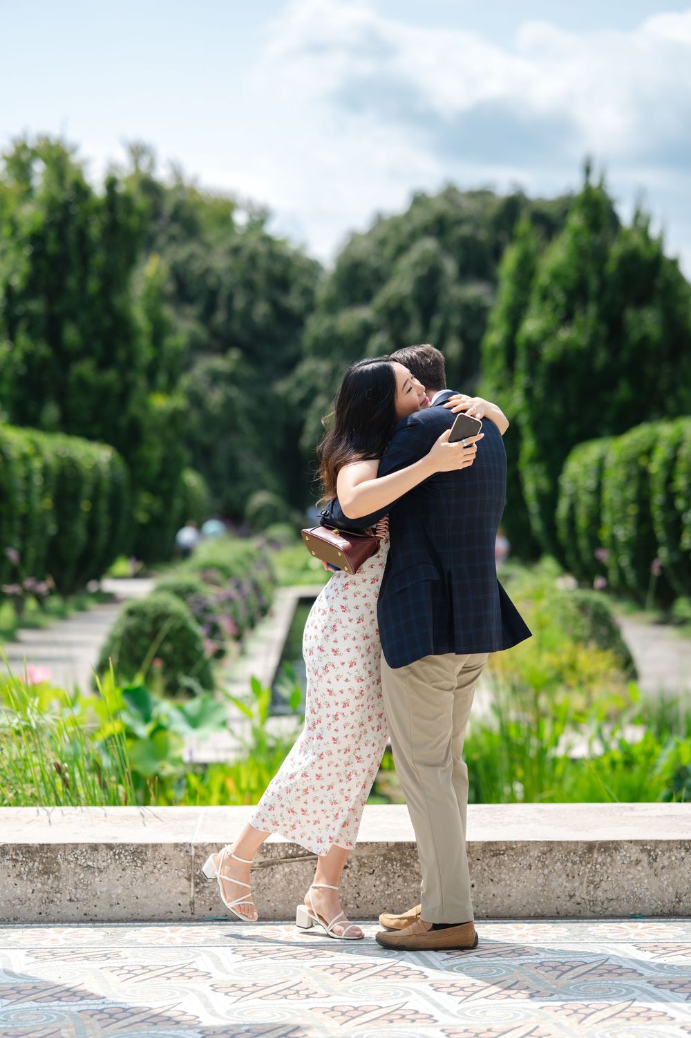 Couple embracing in a lush garden, woman in floral dress and man in navy blazer, sharing a romantic moment outdoors.