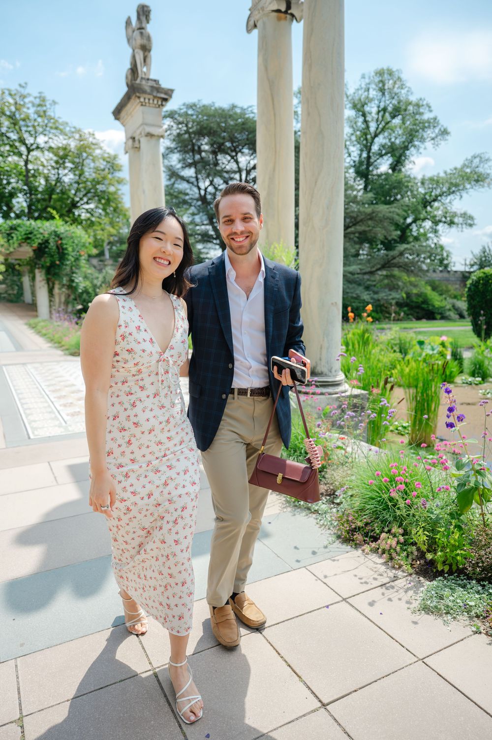 Couple smiling together in a formal garden with stone columns and colorful flowers on a sunny day.