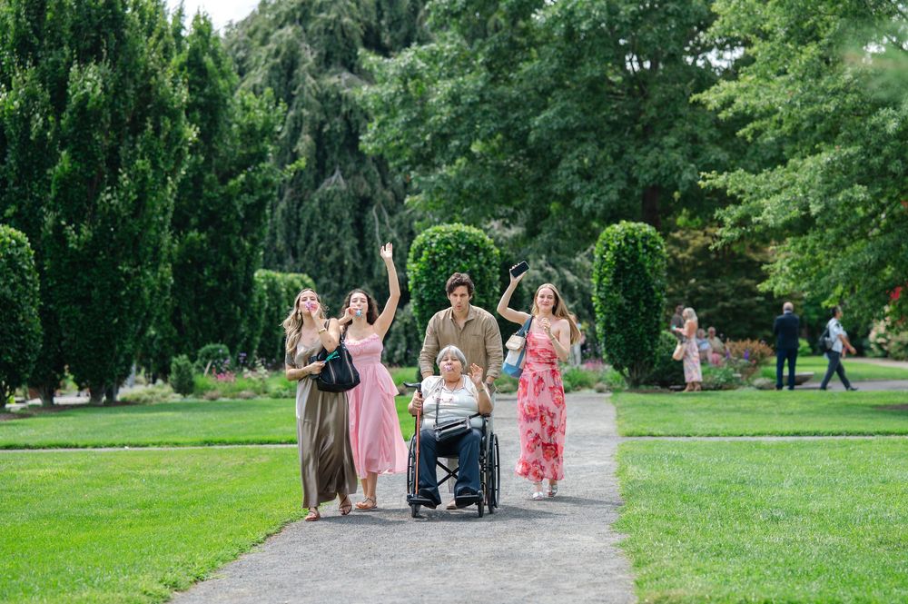 Family group with wheelchair strolling along a scenic garden path surrounded by tall green trees.