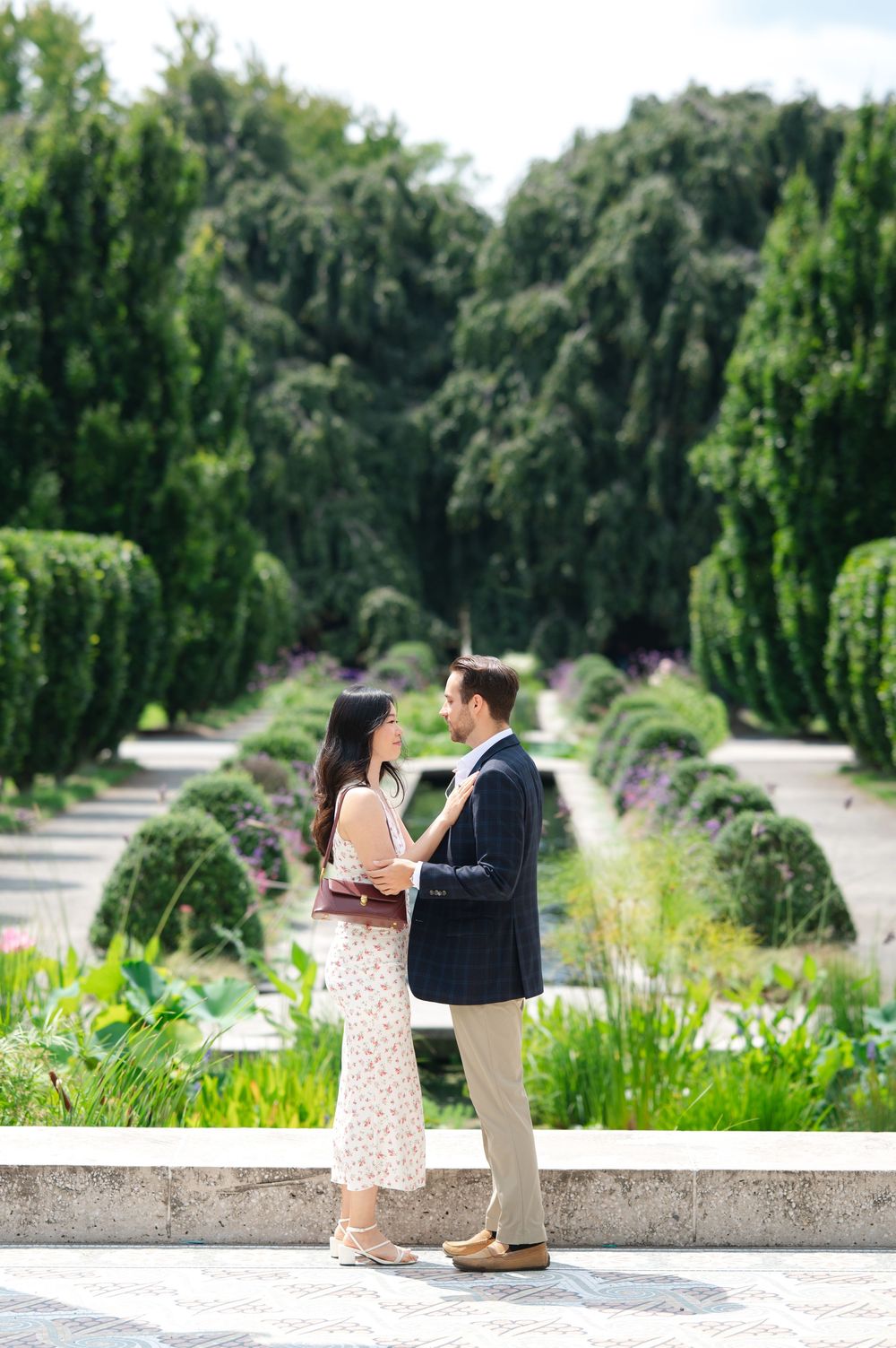 Couple in elegant attire stands facing each other in a lush formal garden with manicured hedges and trees.