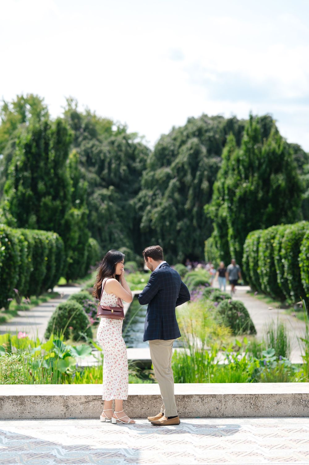 Couple standing face-to-face in a lush garden with manicured hedges and trees in the background.