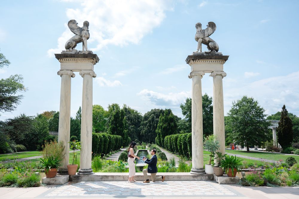 Couple posing between ornate columns topped with griffin statues in a formal garden with lush greenery.