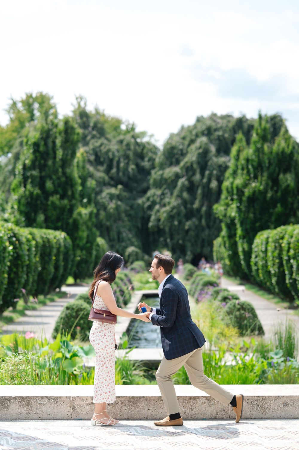 Man kneeling on one knee proposing to woman in a lush green garden setting on a sunny day.