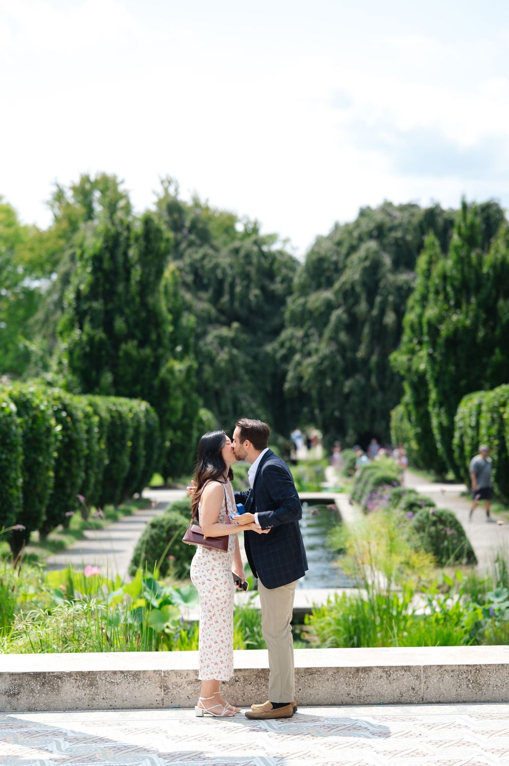 Couple sharing a romantic kiss in a lush formal garden with manicured hedges and a fountain pathway.