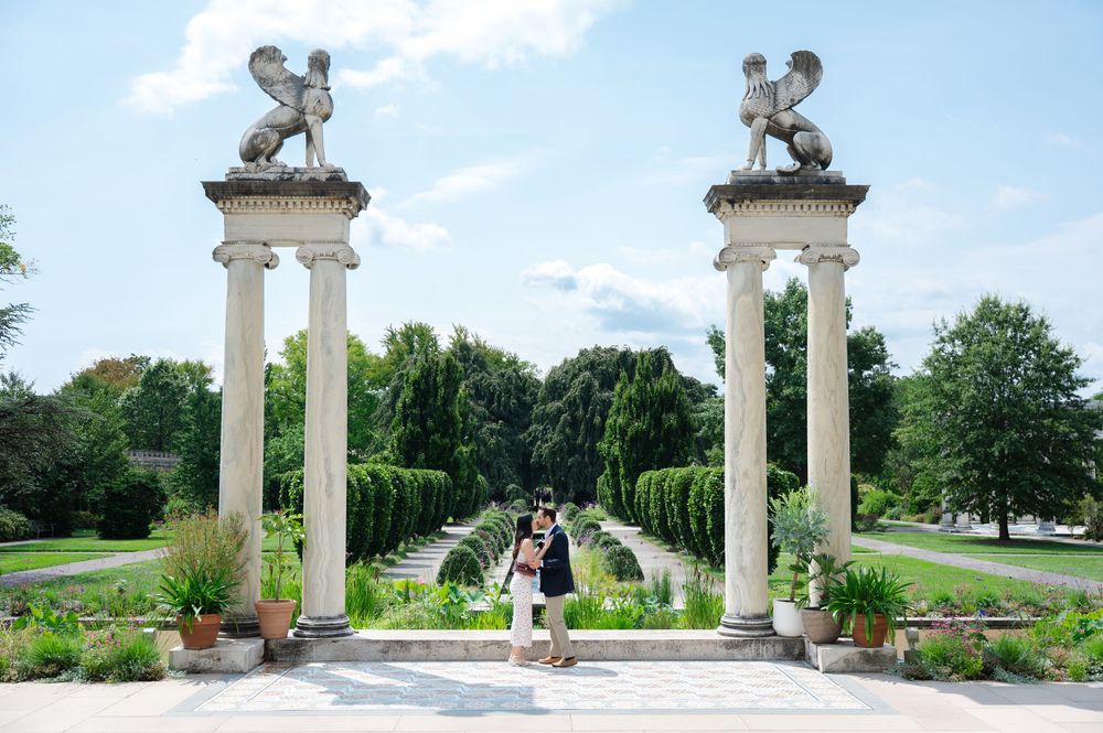 Couple posing between ornate stone columns topped with sphinx statues at a formal garden with fountains.