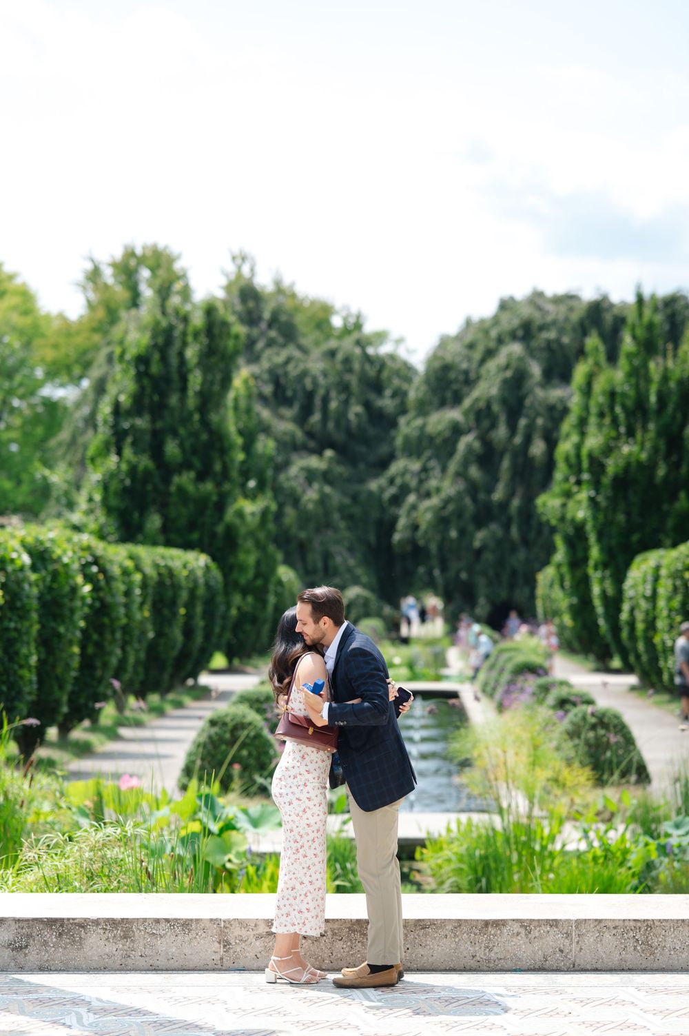 Couple embracing and kissing in a lush formal garden with manicured hedges and a reflecting pool.